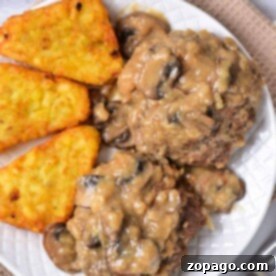 A plate showcasing an old-fashioned Salisbury steak recipe, served with rich mushroom gravy and crispy hash browns, rests elegantly on a beige placemat.
