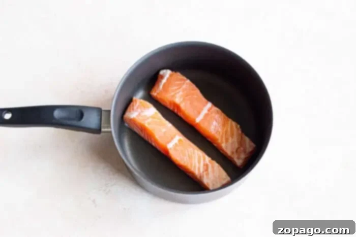Poached Fish 3 Two raw salmon fillets are nestled side by side in a non-stick pan with a black handle, ready to sizzle and infuse flavors on a light-colored surface.