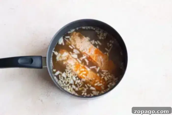 Poached Fish 5 A black saucepan with fish fillets gently boiled in a seasoned broth, topped with chopped onions. The pan rests on a light-colored countertop, promising a delicious and comforting meal.