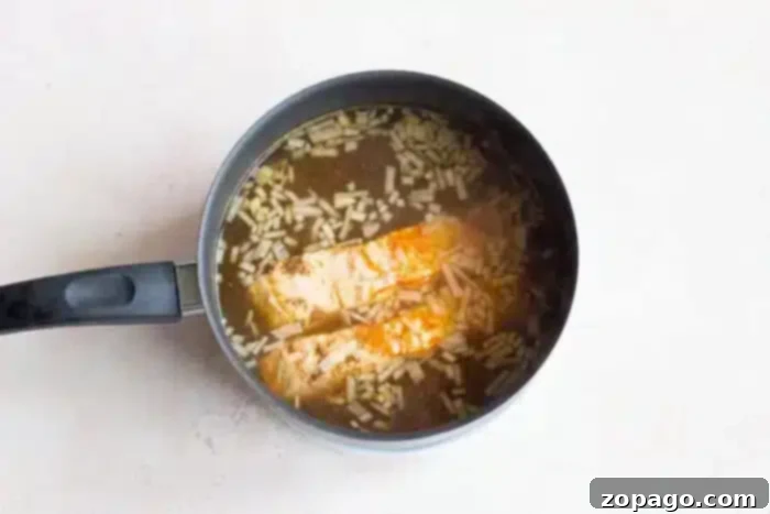 Poached Fish 8 A saucepan on a light surface contains two pieces of boiled fish cooking in a broth with visible rice grains. Steam rises above the mixture, indicating it is being heated.