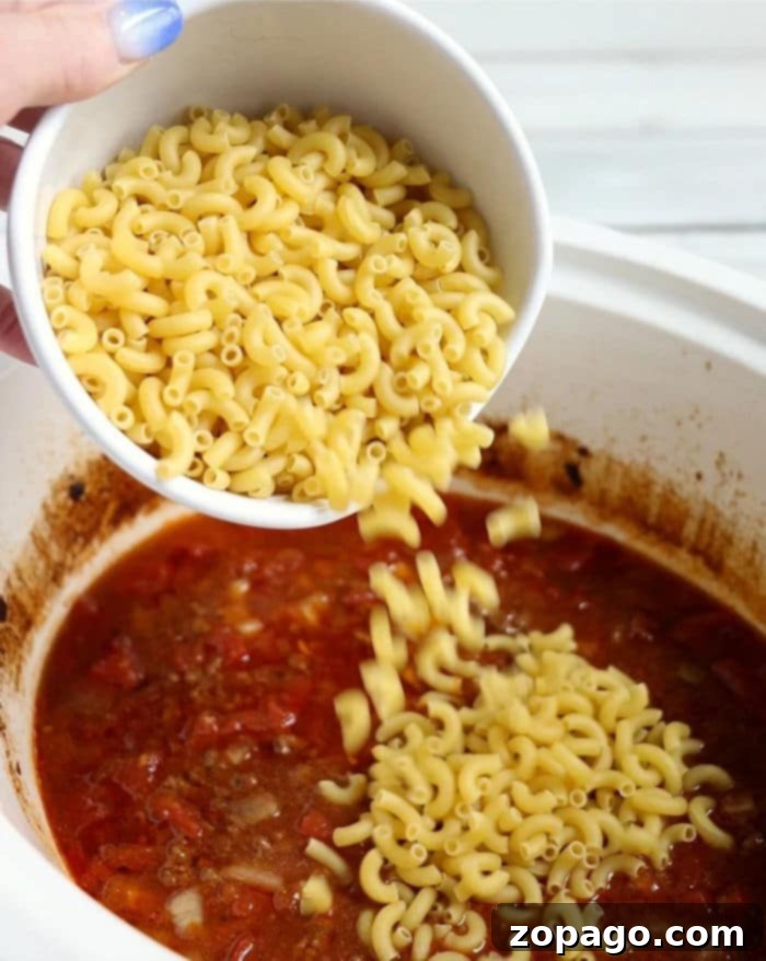 Effortless Slow Cooker Sausage Meals 7 Pouring uncooked elbow macaroni into a simmering pot of crockpot chili mac with a rich tomato base adds hearty texture and delicious flavor.