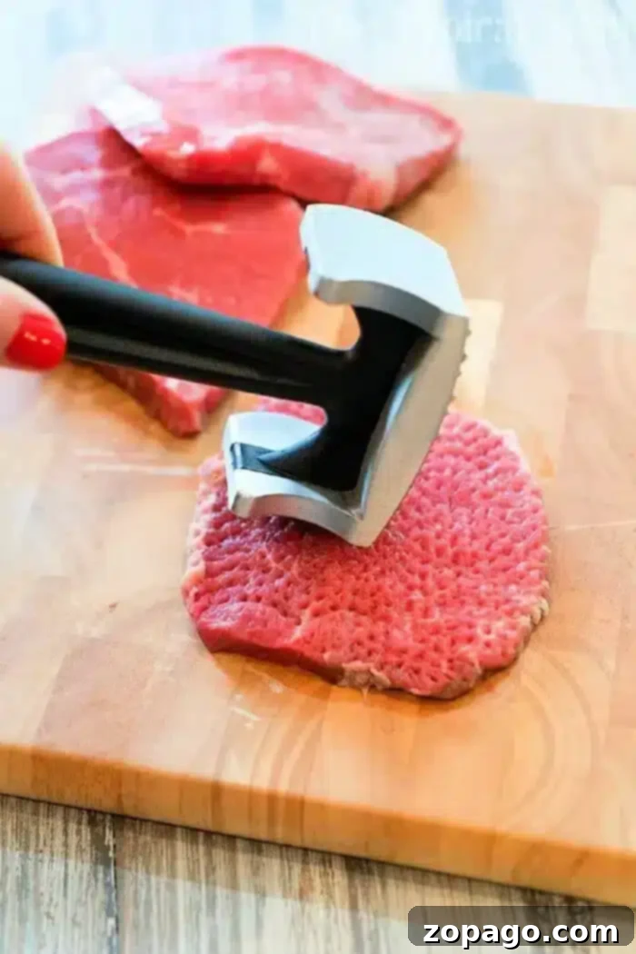 Delicious Minute Steaks in Savory Gravy 4 A person is expertly using a metal meat tenderizer on a piece of raw meat, preparing it for delicious minute steak recipes. The wooden cutting board holds the focus, while additional slices of meat patiently await their turn in the background.