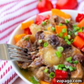 Bowl of easy oxtail stew with carrots, bell peppers, and green onions, with a fork and tomatoes in the background.