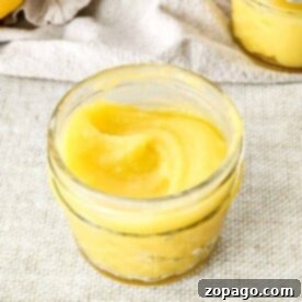 A small glass jar filled with smooth yellow lemon curd rests invitingly on a textured beige surface, enticing thoughts of delectable lemon curd desserts.