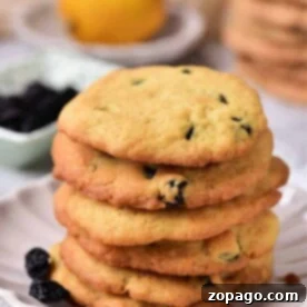 A delightful stack of lemon blueberry cookies rests on a white plate, with a fresh lemon and a vibrant bowl of blueberries adding charm to the background.