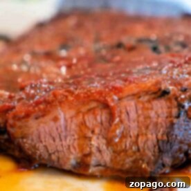 Close-up of a Crockpot Brisket Recipe with a glistening, caramelized exterior. The meat appears juicy and tender, with a portion cut to show its texture. A colorful, blurred background contrasts with the rich, reddish-brown tones of the brisket.