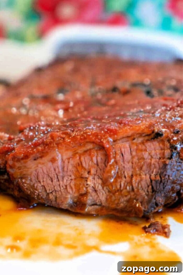 Melt In Your Mouth Slow Cooker Brisket 9 Close-up of a Crockpot Brisket Recipe with a glistening, caramelized exterior. The meat appears juicy and tender, with a portion cut to show its texture. A colorful, blurred background contrasts with the rich, reddish-brown tones of the brisket.