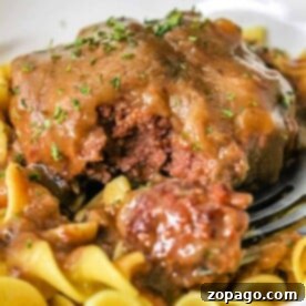 Close-up of beef smothered in brown gravy on egg noodles, reminiscent of a classic chopped steak recipe, garnished with herbs in a white bowl.