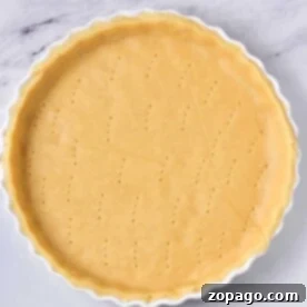 Unbaked pie crust in a white tart pan, pierced with fork holes, rests on a marble surface, ready for the next step in learning how to blind bake a pie crust.