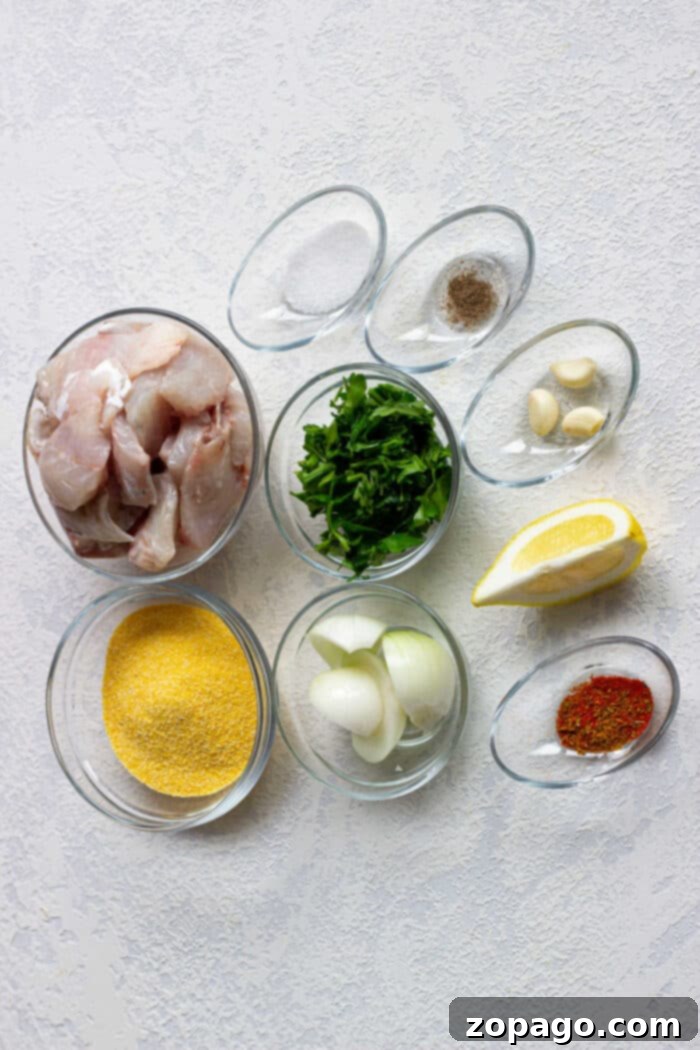 Ingredients for a dish: raw cod, herbs, lemon wedge, onion, spices, garlic, and cornmeal in small bowls. Prepare this mix with the same care you would use for crafting delicate Cod Fish Cakes.