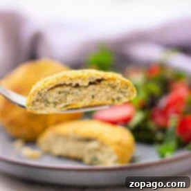 Close-up of a fork holding a breaded delicious cod fish cakes. A salad is blurred in the background on a gray plate.