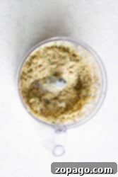 A food processor containing a coarse, creamy mixture of herbs and spices sits on a light countertop, ready to be transformed into delicious cod fish cakes.