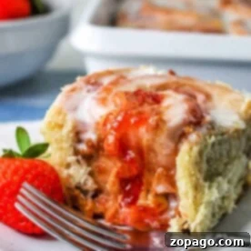 Sweet Peanut Butter Rolls 8 A peanut butter sweet roll with strawberry filling is topped with icing and sits on a white plate. A fork rests nearby, accompanied by a fresh strawberry. In the background, more sweet rolls can be seen in a baking dish.