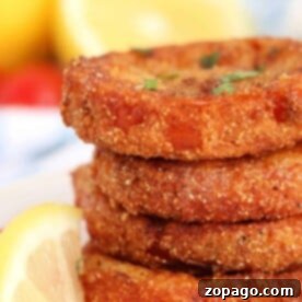 Stacked crispy fried red tomatoes are garnished with fresh herbs, served alongside lemon wedges in the background.