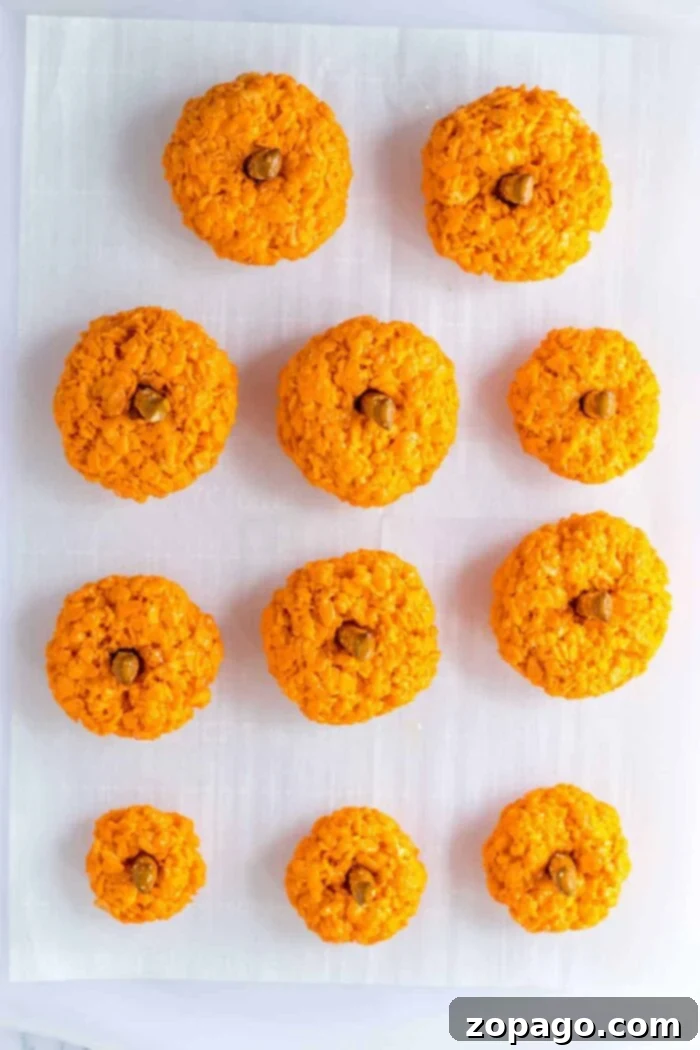 Crispy Autumn Pumpkin Bites 5 Ten Halloween Rice Krispie Treats, charmingly shaped like pumpkins with green stems and vines, arranged in rows on white parchment paper.
