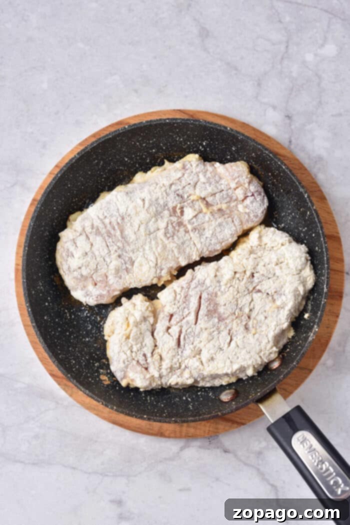 Pork Cutlets 4 Two flour-coated pork steaks sizzle alongside a perfectly seasoned pork cube steak in a black frying pan on a marble surface.