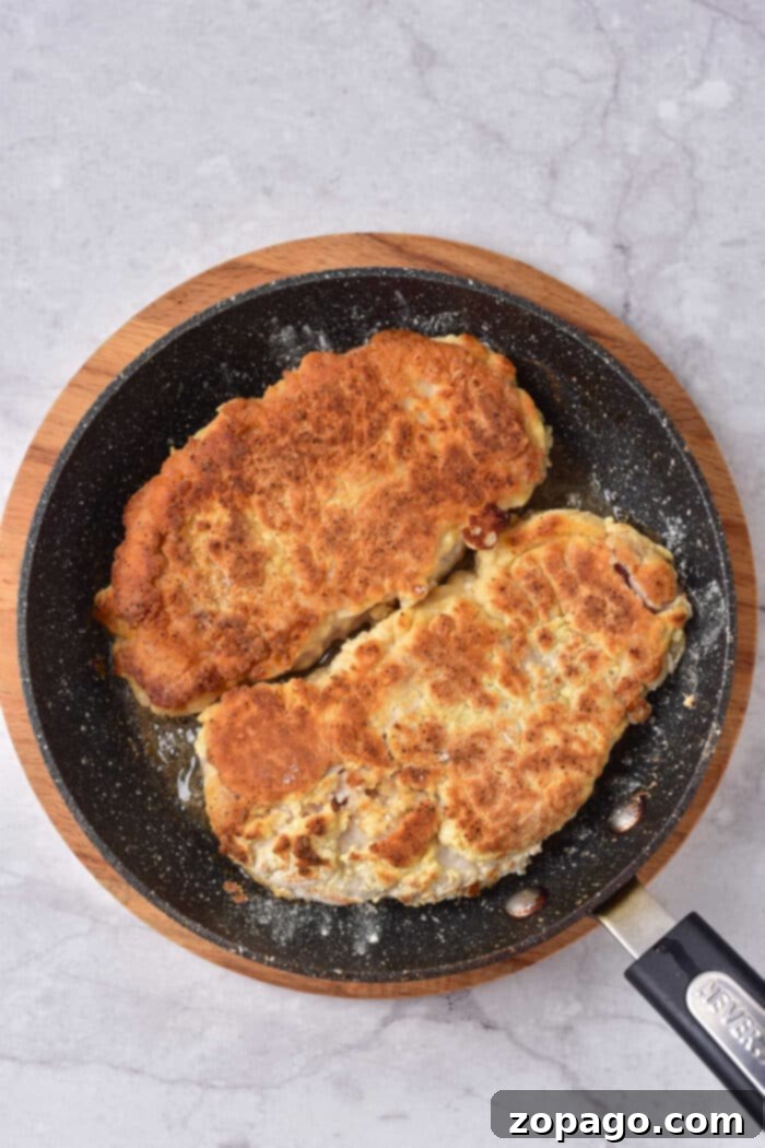 Two breaded pork cutlets and a succulent pork cube steak sizzle in a black pan set on a wooden board.