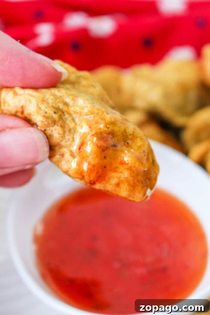 Crispy Tempura Chicken Bites 2 Close-up of a hand holding a golden-brown tempura chicken nuggets, dipped in a bowl of red sauce. A red cloth adds warmth to the background.