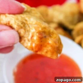 Crispy Tempura Chicken Bites 4 Close-up of a hand holding a golden-brown tempura chicken nugget, dipped in a bowl of red sauce. A red cloth adds warmth to the background.