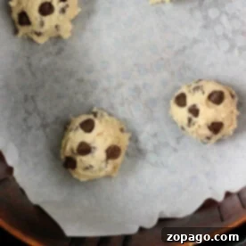 Air Fryer Cookie Dough Perfection 4 Four air fryer cookie dough balls arranged on a parchment-lined baking sheet. The dough balls are spaced apart, ready for baking. The bottom edge of a circular container is visible at the bottom of the image.