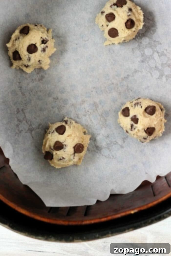 Air Fryer Cookie Dough Perfection 7 Four air fryer cookie dough balls arranged on a parchment-lined baking sheet. The dough balls are spaced apart, ready for baking. The bottom edge of a circular container is visible at the bottom of the image.