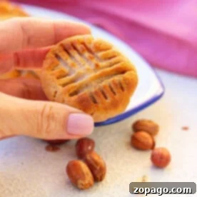 Hand holding an air fryer peanut butter cookie with a classic fork design, surrounded by peanuts on a light surface.