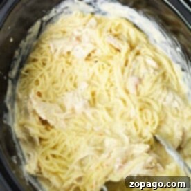 Angel Crockpot Chicken: Creamy pasta with shredded chicken in a black slow cooker, elegantly served with metal tongs.