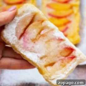 A hand delicately holds a flaky peach puff pastry dusted with powdered sugar, with more of these tempting pastries in the background.