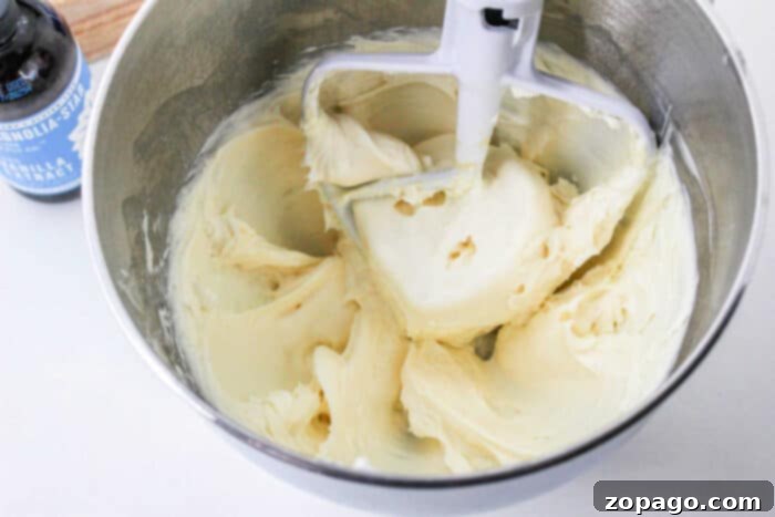 Smooth Butter-Free Cream Cheese Frosting 4 Creamy frosting in a mixing bowl with a paddle attachment, next to a bottle of vanilla extract, promises a rich flavor. Consider topping it with smooth cream cheese frosting without butter for an indulgent finish.