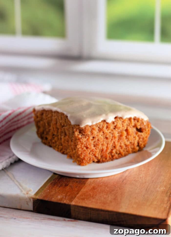 Simple Pumpkin Magic Cake 5 A slice of frosted Crazy Cake carrot cake sits on a white plate. The cake has a thick, soft texture and is topped with a light layer of white icing. The plate rests on a wooden board, with a white and red striped cloth partially visible in the background.