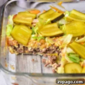 Glass dish with a layered Big Mac Casserole, topped with sliced pickles and crisp lettuce, one mouthwatering portion removed.