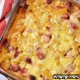 Cheesy Hot Dog Casserole Bake 7 A baked dish in a glass pan, this hot dog casserole boasts a golden crust with visible slices of sausages and pasta, possibly macaroni. A red and white cloth is partially visible on the side.