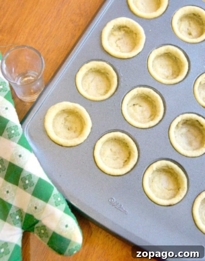 Cookie Cups: Tiny Treats, Big Flavor 2 Mini cookie cups filled with various toppings, neatly arranged in a mini muffin pan beside a green checkered oven mitt and a small glass on a rustic wooden surface. The image highlights their miniature size and delicious appeal.