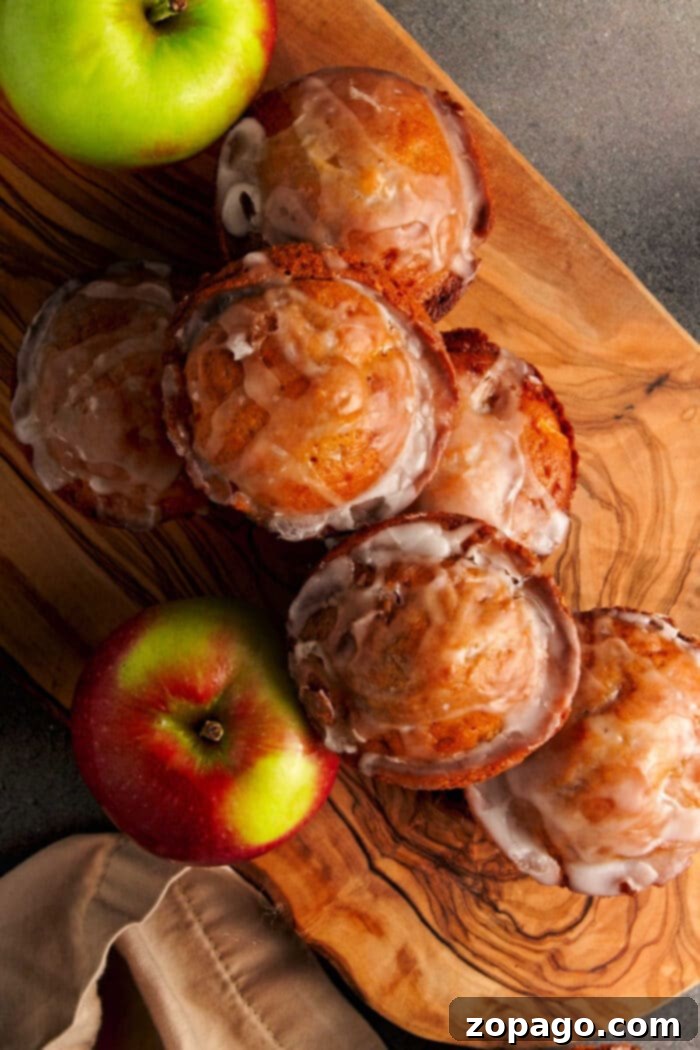 Sweet & Spiced Apple Fritter Muffins 2 Exquisitely baked apple fritter muffins, glistening with a sweet glaze, are arranged on a rustic wooden surface. Two fresh apples, one green and one red, complete the inviting autumn scene, perfectly capturing the essence of baked apple fritters.