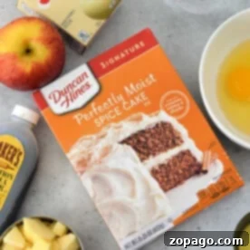 An array of tempting ingredients awaits on the kitchen counter: a box for Apple Cake with Cake Mix, creamy pudding, aromatic cinnamon, chopped apples, an egg, golden syrup, and a trusty measuring cup to tie it all together.