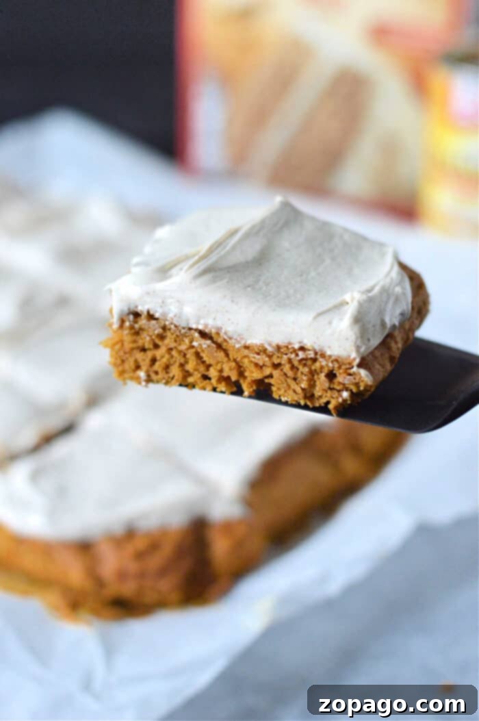 Easy Cake Mix Pumpkin Bars 6 A frosted square pumpkin bars with cake mix, is lifted on a spatula above more slices on parchment paper.