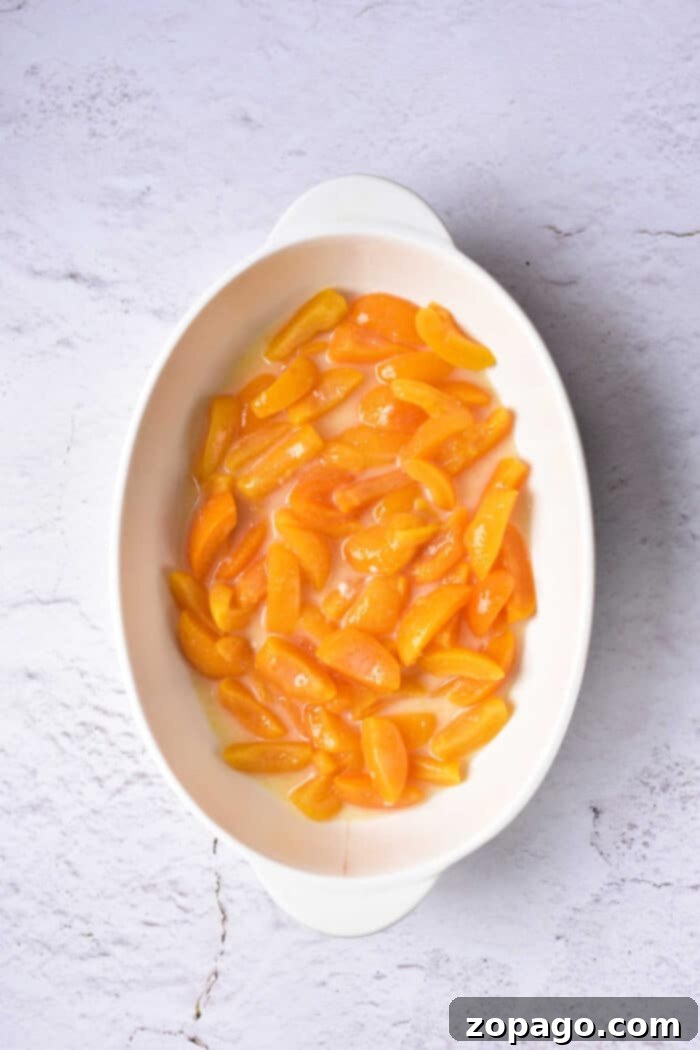 Oval dish with sliced apricots in syrup, reminiscent of an apricot cobbler, placed on a textured white surface.
