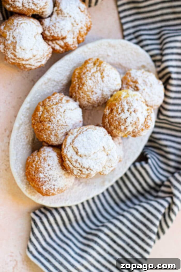 Crispy Biscuit Donut Bites 7 A white oval plate holds several powdered sugar-covered cream puffs, resembling delicate biscuit donut holes. More cream puffs are partially visible in the top left corner. The plate is placed on a light pink surface next to a black and white striped cloth.