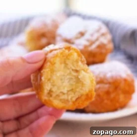 A hand delicately holds a partially eaten fried biscuit donut hole, with a plate piled high with similar treats tempting in the background.
