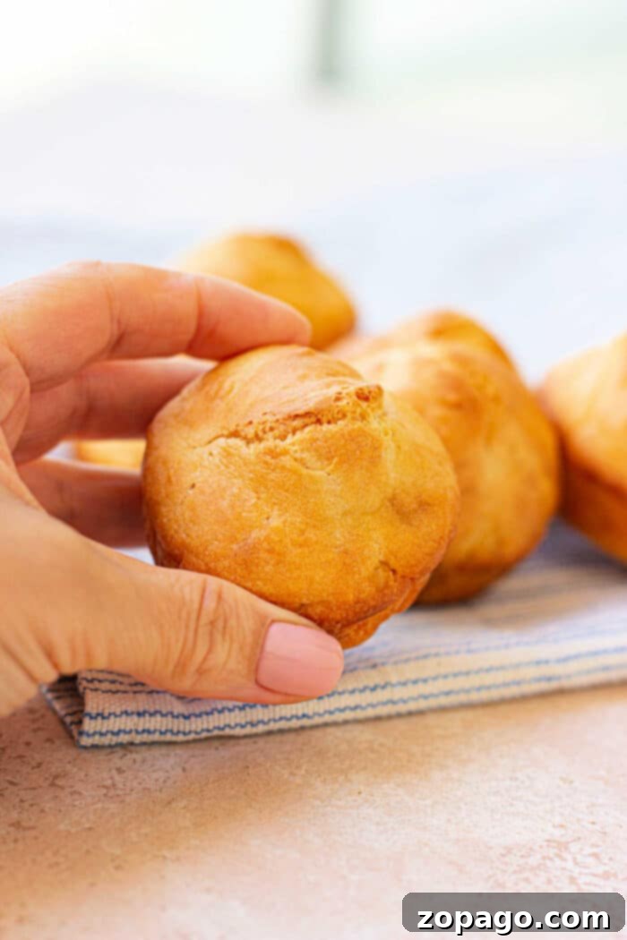 Fluffy Mayonnaise Biscuits 5 A hand holding a freshly baked mayo biscuit over a striped cloth resembles the warmth of homemade mayonnaise biscuits, with more muffins tempting in the background.