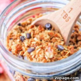 A jar of homemade granola with chocolate chips and a wooden tablespoon inside, strawberries temptingly arranged in the background.
