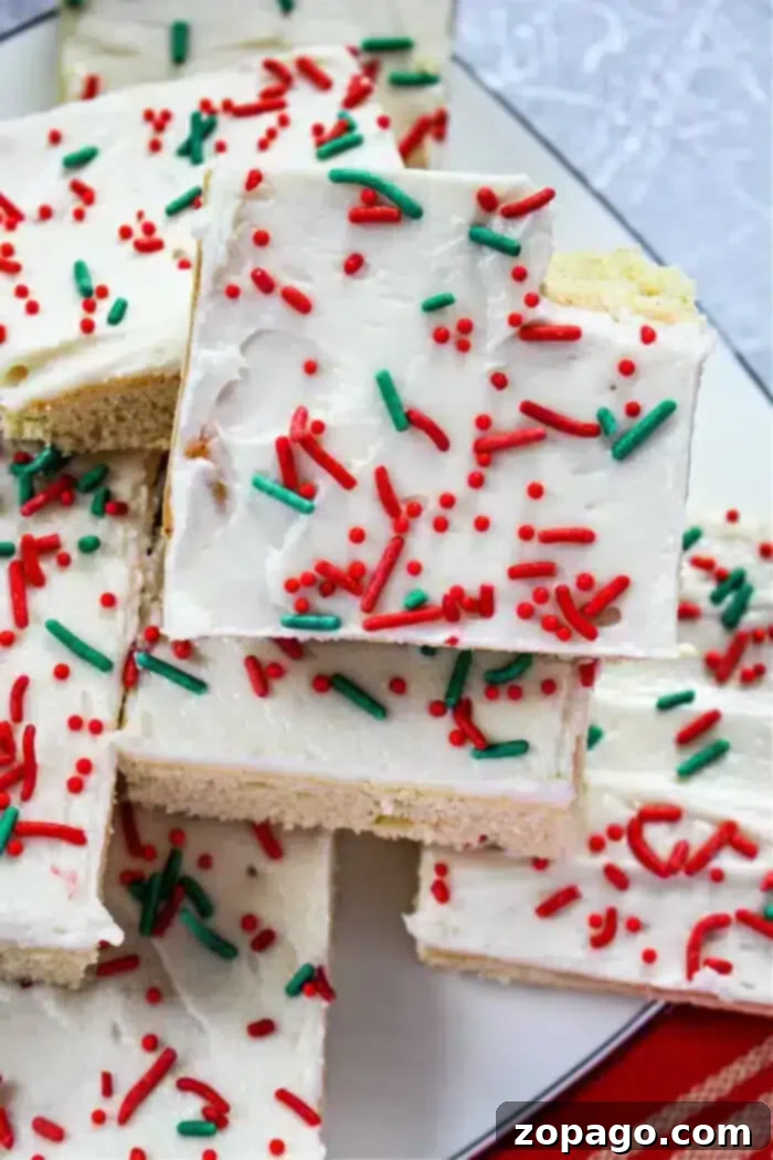 Easy Sugar Cookie Bars 2 A white plate holds a stack of frosted Christmas bar cookies. The cookies are topped with white frosting and decorated with red and green sprinkles. There is a red and white striped cloth partially visible at the bottom.
