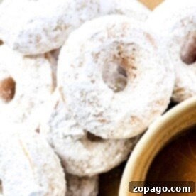 Whip Up Powdered Sugar Donuts 8 A close-up of several powdered sugar donuts stacked on a plate, with a cup filled with a rich, brown liquid, likely coffee or tea.