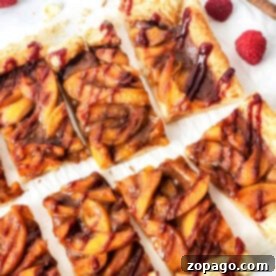 Sliced peach puff pastry tart with a flaky crust, drizzled with raspberry sauce, surrounded by fresh raspberries on parchment paper.