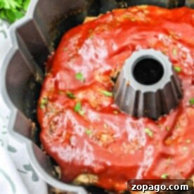 Meatloaf Ring 6 A Bundt Pan Meatloaf glistens under a glossy tomato sauce, with fresh parsley leaves artfully sprinkled on top. In the background, a bunch of parsley rests beside a patterned cloth, complementing this culinary centerpiece.