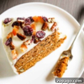 A slice of sweet potato cake with cream cheese frosting, pecans, and dried fruit sits on a white plate with a fork.