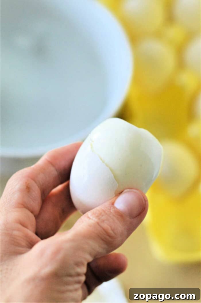 Easy Baked Hard Boiled Eggs Toaster Oven Recipe 6 A hand holding a partially peeled boiled egg. In the background, there is a white bowl and a yellow egg carton containing several boiled eggs, hinting they might have been cooked using an eggs in toaster oven method.