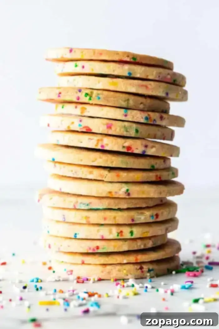 Confetti Sugar Cookie Delights 2 A stack of round Sprinkle Sugar Cookies with colorful sprinkles embedded throughout is displayed against a plain white background. Additional sprinkles are scattered at the base of the stack.