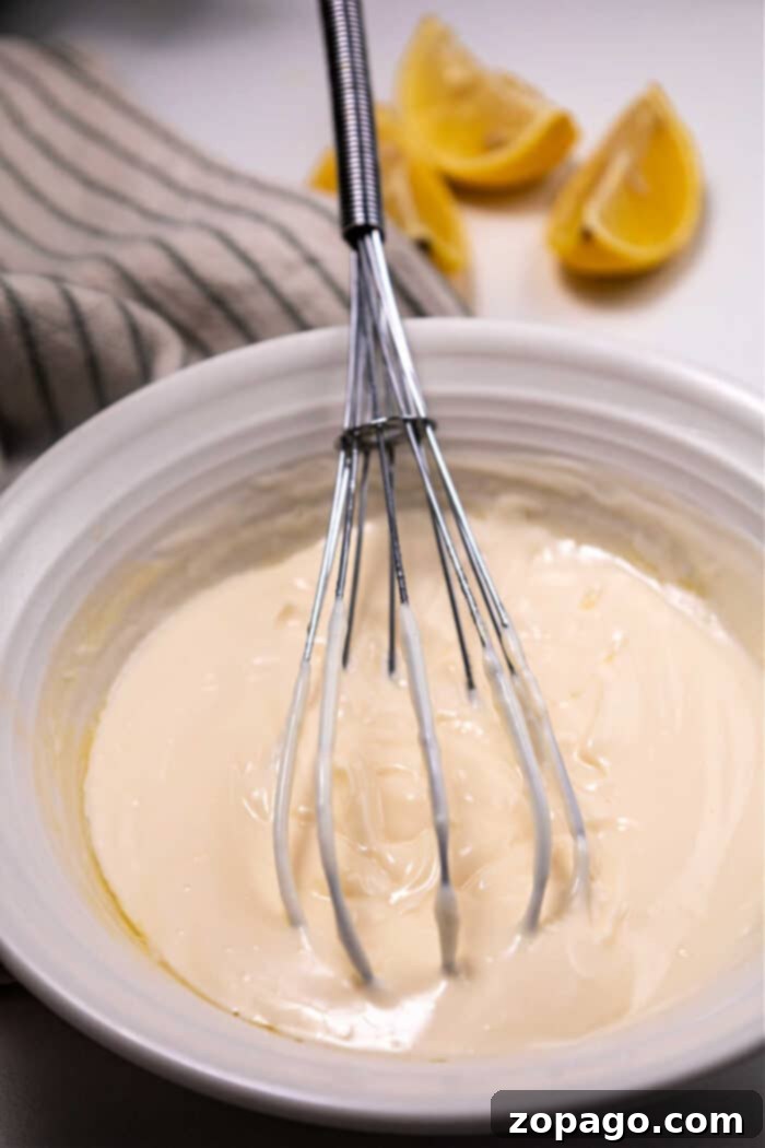 A whisk in a bowl of thick mayonnaise, with lemon wedges and a striped cloth in the background—its almost reminiscent of preparing Mexican mayonnaise.