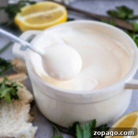 A spoon dips into a white ceramic bowl of creamy Mexican mayonnaise, surrounded by lemon slices and parsley.